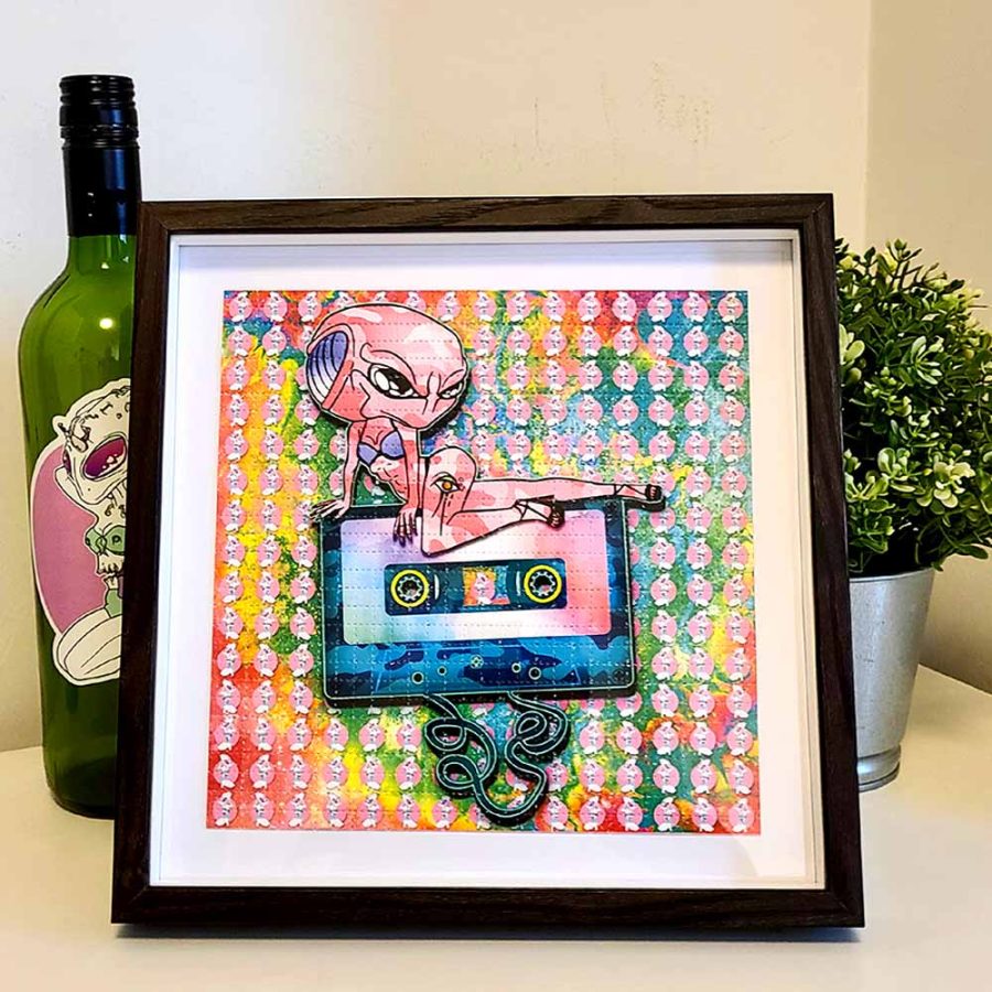 photo of a framed lsd blotter acid art print featuring psychedelic rainbow colours and an alien posing on top of a audio cassette tape trippy gift