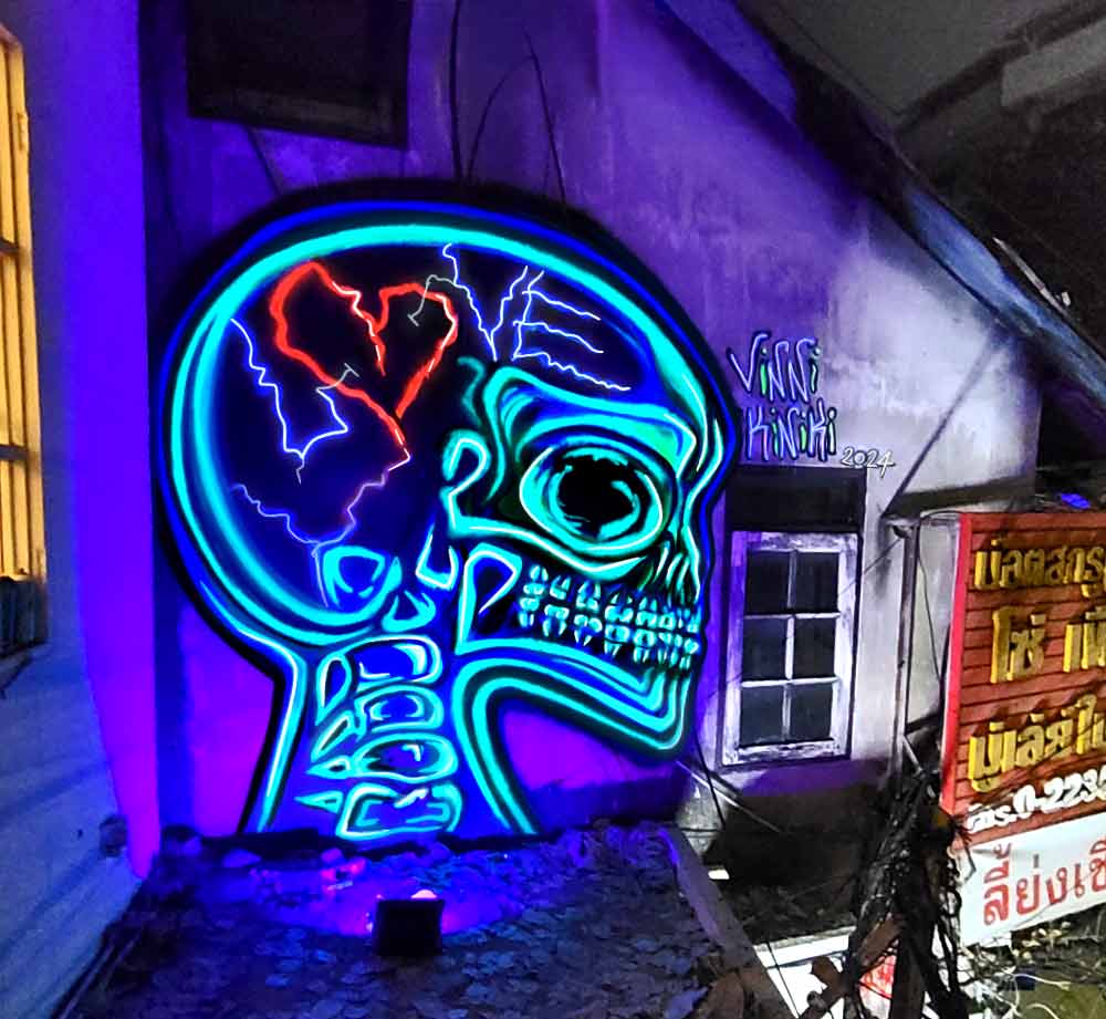 Black light graffiti mural by Vinni Kiniki featuring humanoid alien skull with cranial sutures spelling the word love vibrant neon painted in bangkok, Thailand