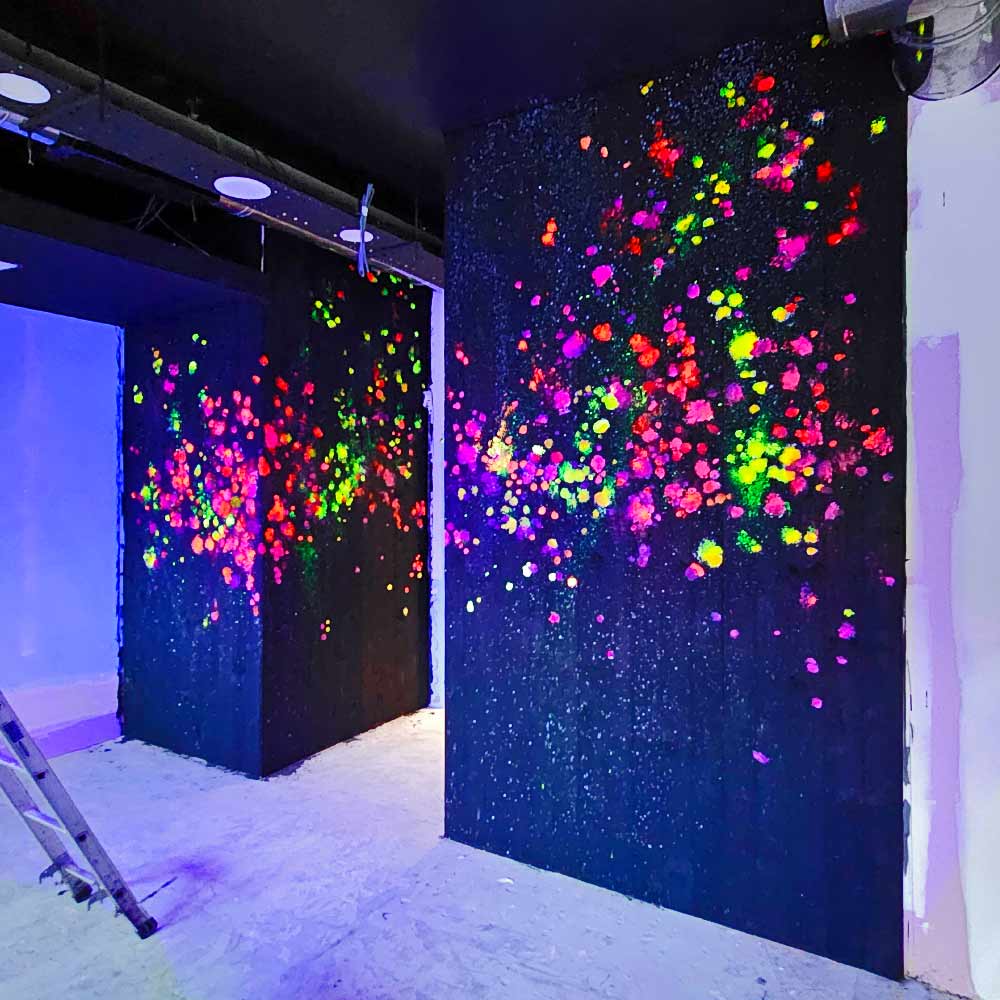 Black light art on dark-stained timber wall at My Thai restaurant in Manchester Piccadilly, painted by artist ViNNi KiNiKi with a UV reactive abstract impressionist mural using brushes and acrylics, creating a textured contrast against the surrounding pop art graffiti.
