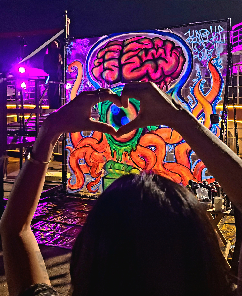 Heart hands held over black light graffiti mural of a psychedelic jellyfish sea monster by ViNNi KiNiKi, painted live on Bangkok Island during Galleries’ Nights Bangkok 2025