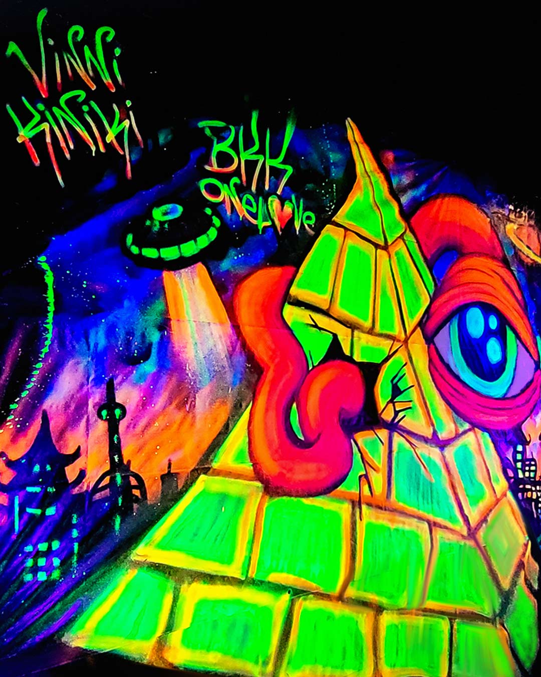 Low-angle shot of fluorescent UV graffiti artwork showing a glowing green pyramid, UFO beam, and skyline painted in Bangkok by ViNNi KiNiKi.