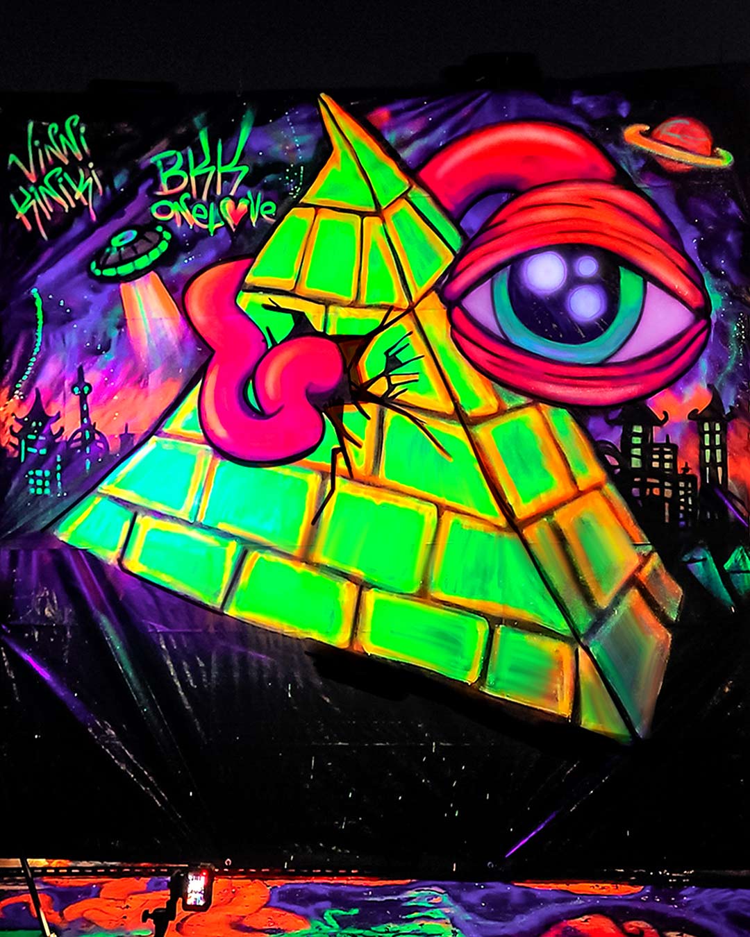 Neon UV blacklight graffiti mural of an Illuminati pyramid and all-seeing eye painted live in Bangkok by artist ViNNi KiNiKi.