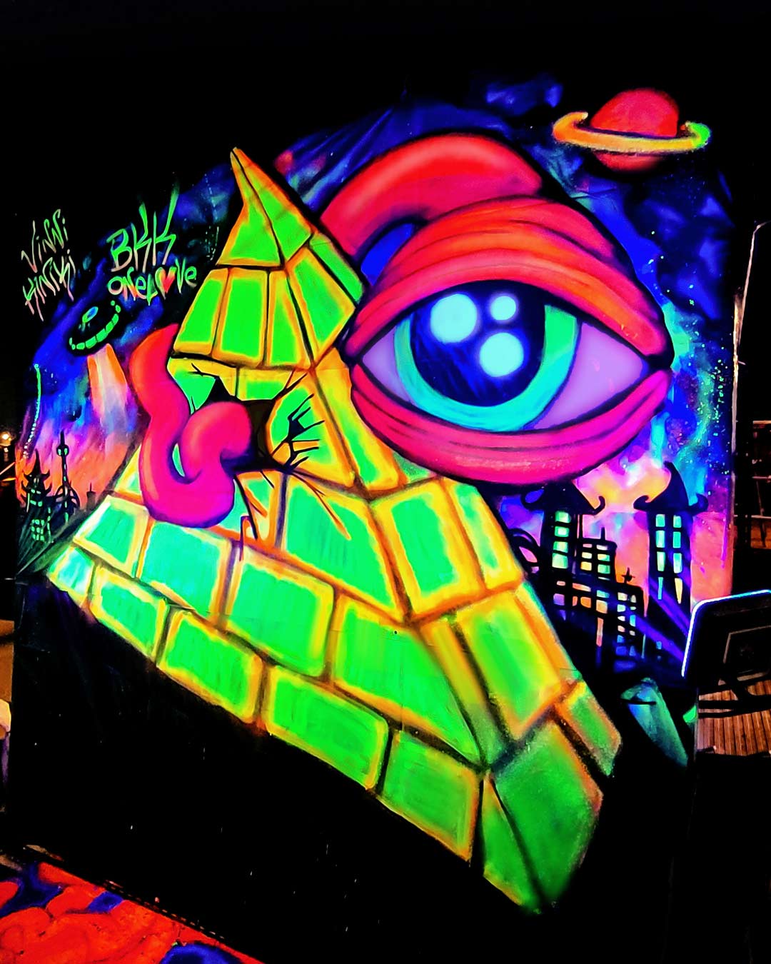 Side perspective of a glowing neon blacklight street art mural featuring a pyramid and psychedelic eye by ViNNi KiNiKi.
